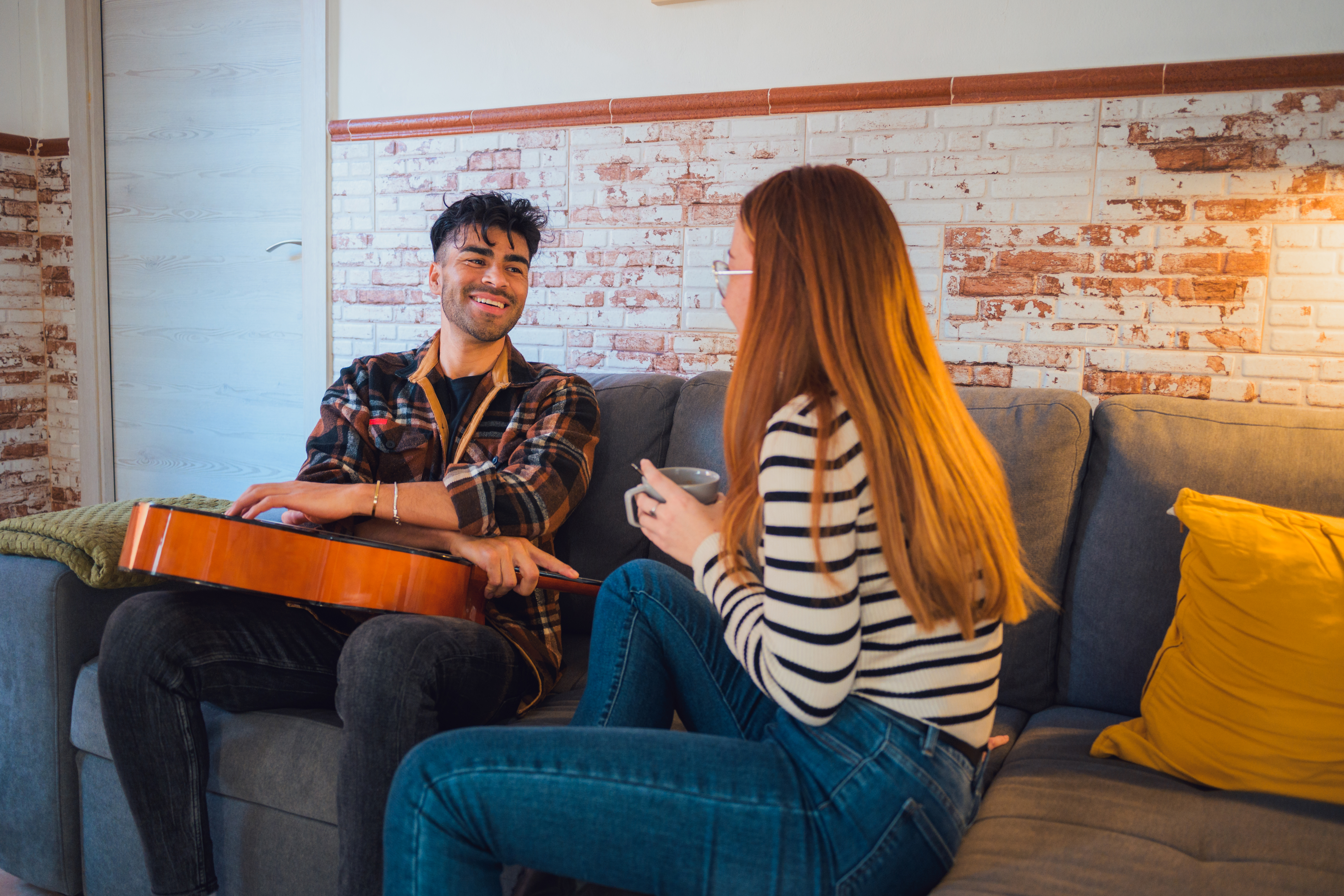 A man and woman are seated on a couch talking with a guitar. They have a good conversation while smiling